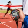 Chronomètre sport numérique AnyTime D309 utilisé par un juge lors d’une course d’athlétisme — écran LCD affichant les temps, boutons rouges « START/STOP » et « SPLIT/LAP », avec lanière noire. Parfait pour le timing précis en compétition ou entraînement.