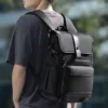 Homme portant un sac à dos business noir imperméable avec rabat et boucle de fermeture, vue arrière montrant le confort des bretelles et le design moderne avec multiples compartiments — idéal pour le travail, les voyages et ordinateur portable.