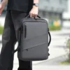 Homme tenant un sac à dos voyage gris anthracite par sa poignée supérieure, design moderne avec bandes diagonales, idéal pour déplacements urbains ou voyages courts — compatible charge USB intégrée.