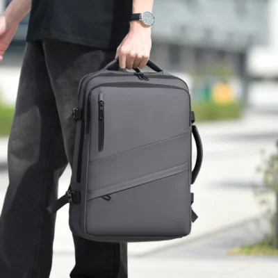Homme tenant un sac à dos voyage gris anthracite par sa poignée supérieure, design moderne avec bandes diagonales, idéal pour déplacements urbains ou voyages courts — compatible charge USB intégrée.