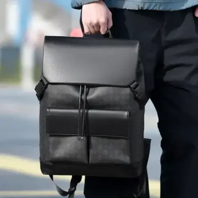Homme tenant le sac à dos business noir à motif monogramme par sa poignée supérieure en extérieur, mettant en valeur la grande capacité, le style urbain et la praticité pour les déplacements professionnels.