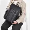Homme assis tenant le sac à dos ordinateur noir à motif monogramme dans les bras, montrant la poignée supérieure et le design vintage élégant — sac imperméable et robuste parfait pour le travail, les études et les déplacements urbains.