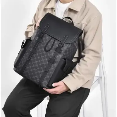 Homme assis tenant le sac à dos ordinateur noir à motif monogramme dans les bras, montrant la poignée supérieure et le design vintage élégant — sac imperméable et robuste parfait pour le travail, les études et les déplacements urbains.