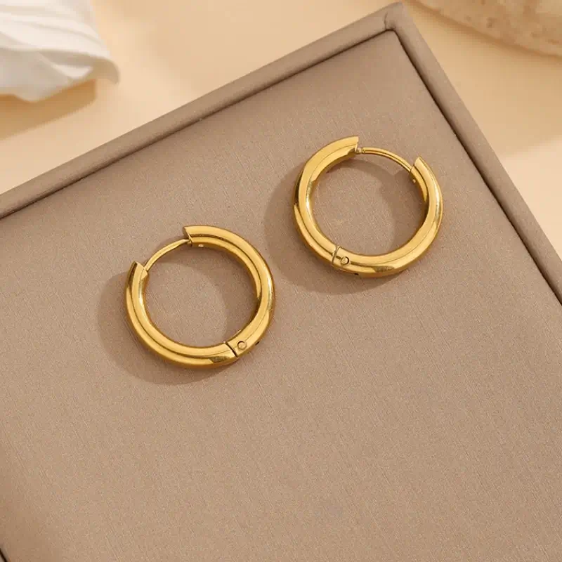 Boucles d'oreilles créoles dorées anti-ternissement avec fermoir à charnière sécurisé, design circulaire épuré et finition brillante — accessoires de mode intemporels et raffinés pour femme, présentés sur un coffret beige.