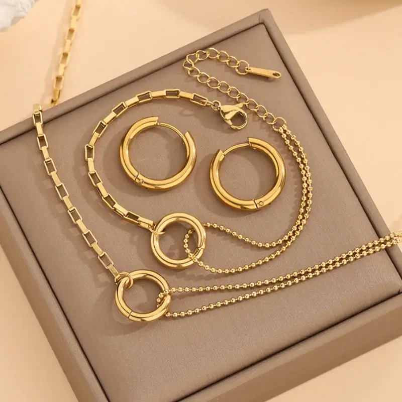 Parure complète 3 pièces dorée anti-ternissement comprenant collier à double chaîne, bracelet assorti et boucles d'oreilles créoles, tous ornés d'un motif anneau circulaire — set de bijoux femme élégant présenté sur écrin, parfait pour un cadeau.