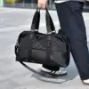 Sac de voyage à main noir haute gamme pour homme, design élégant combinant tissu à motif discret et base en simili-cuir robuste, idéal pour les week-ends ou le sport, montré porté à la main avec sa bandoulière amovible.