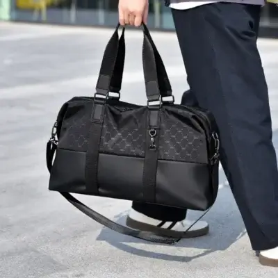 Sac de voyage à main noir haute gamme pour homme, design élégant combinant tissu à motif discret et base en simili-cuir robuste, idéal pour les week-ends ou le sport, montré porté à la main avec sa bandoulière amovible.