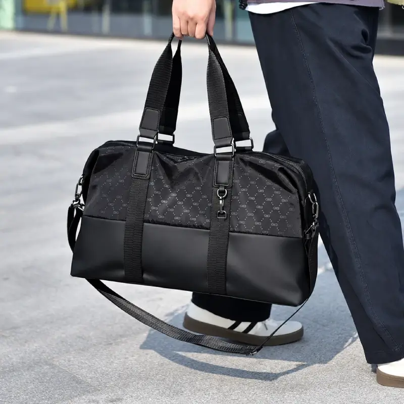 Sac de voyage à main noir haute gamme pour homme, design élégant combinant tissu à motif discret et base en simili-cuir robuste, idéal pour les week-ends ou le sport, montré porté à la main avec sa bandoulière amovible.
